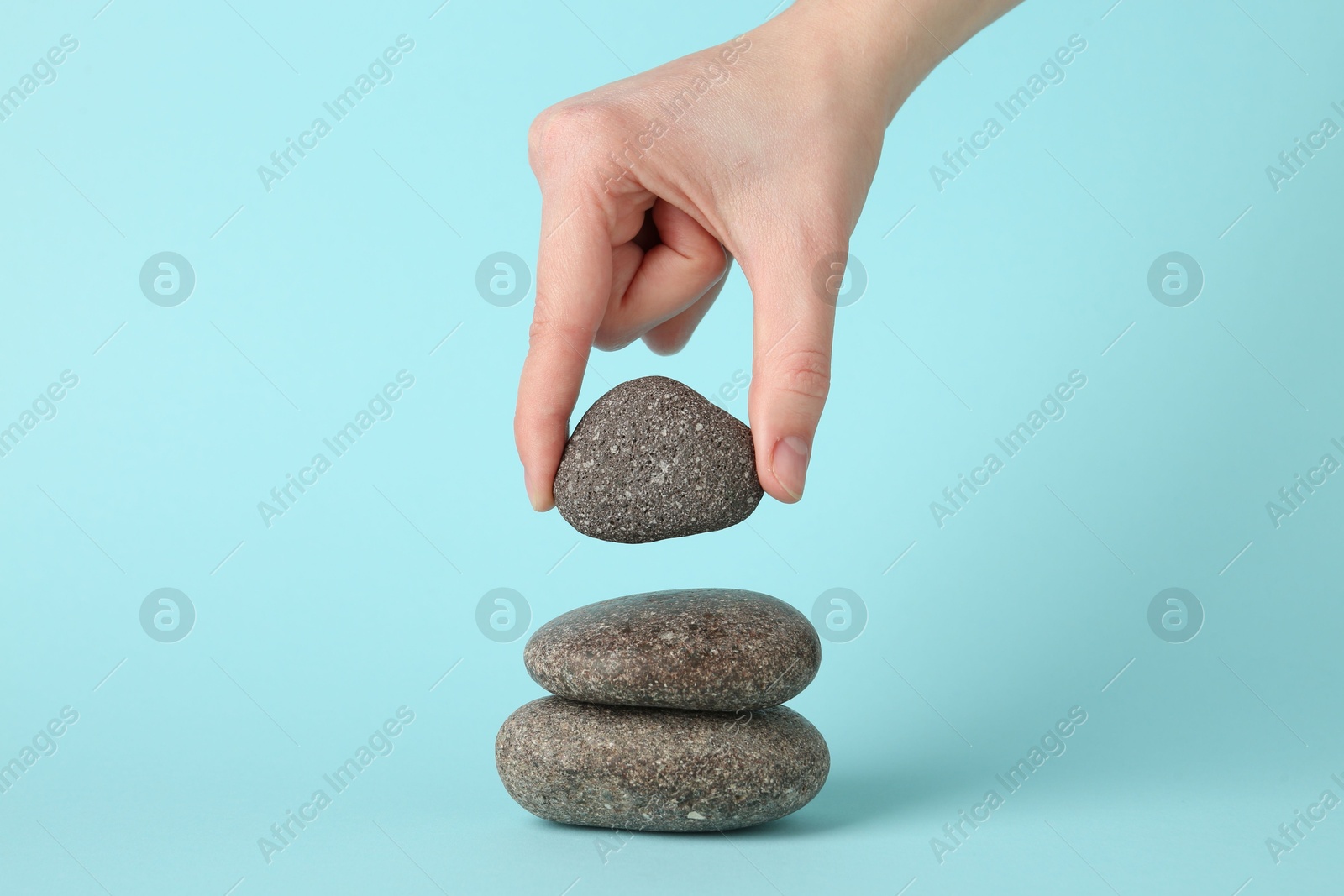 Photo of Woman making stack of stones on light blue background, closeup. Harmony and life balance