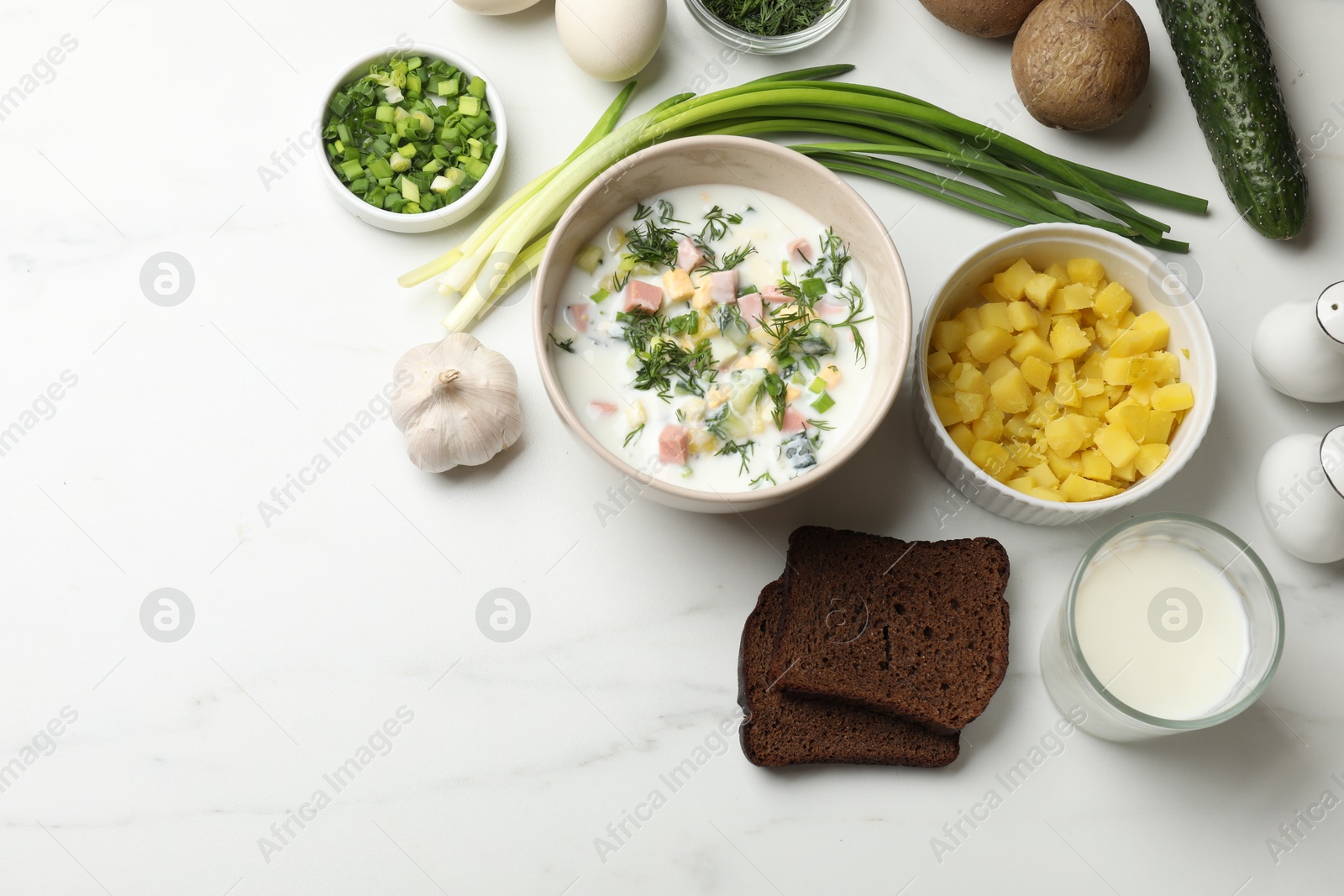 Photo of Delicious okroshka soup in bowl, garlic, bread and ingredients on white marble table, flat lay. Space for text