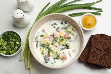 Photo of Delicious okroshka soup in bowl, green onion, bread and spices on white marble table, flat lay