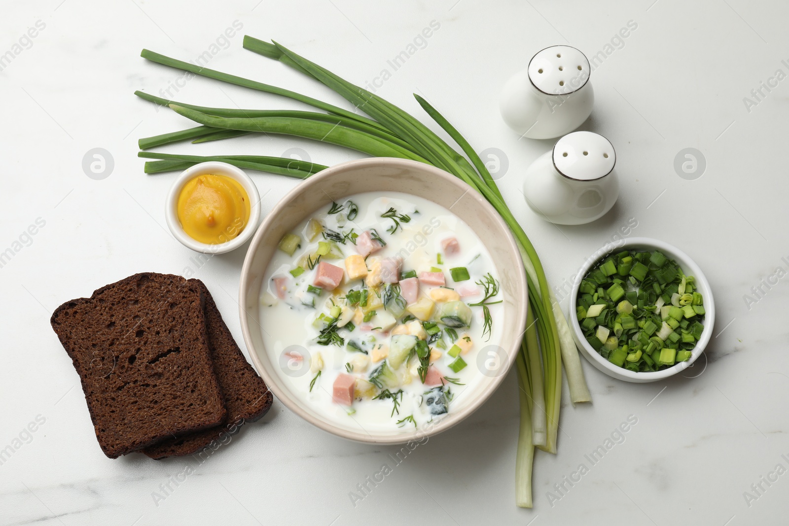 Photo of Delicious okroshka soup in bowl, green onion, bread and spices on white marble table, flat lay