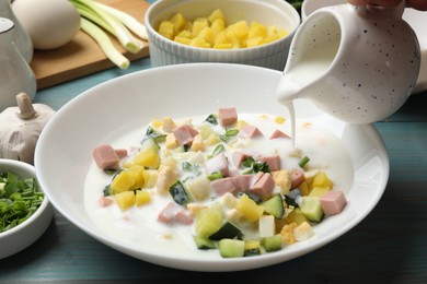 Photo of Making okroshka soup. Pouring kefir into bowl with different ingredients at light blue wooden table, closeup