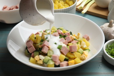 Photo of Making okroshka soup. Pouring kefir into bowl with different ingredients at light blue wooden table, closeup