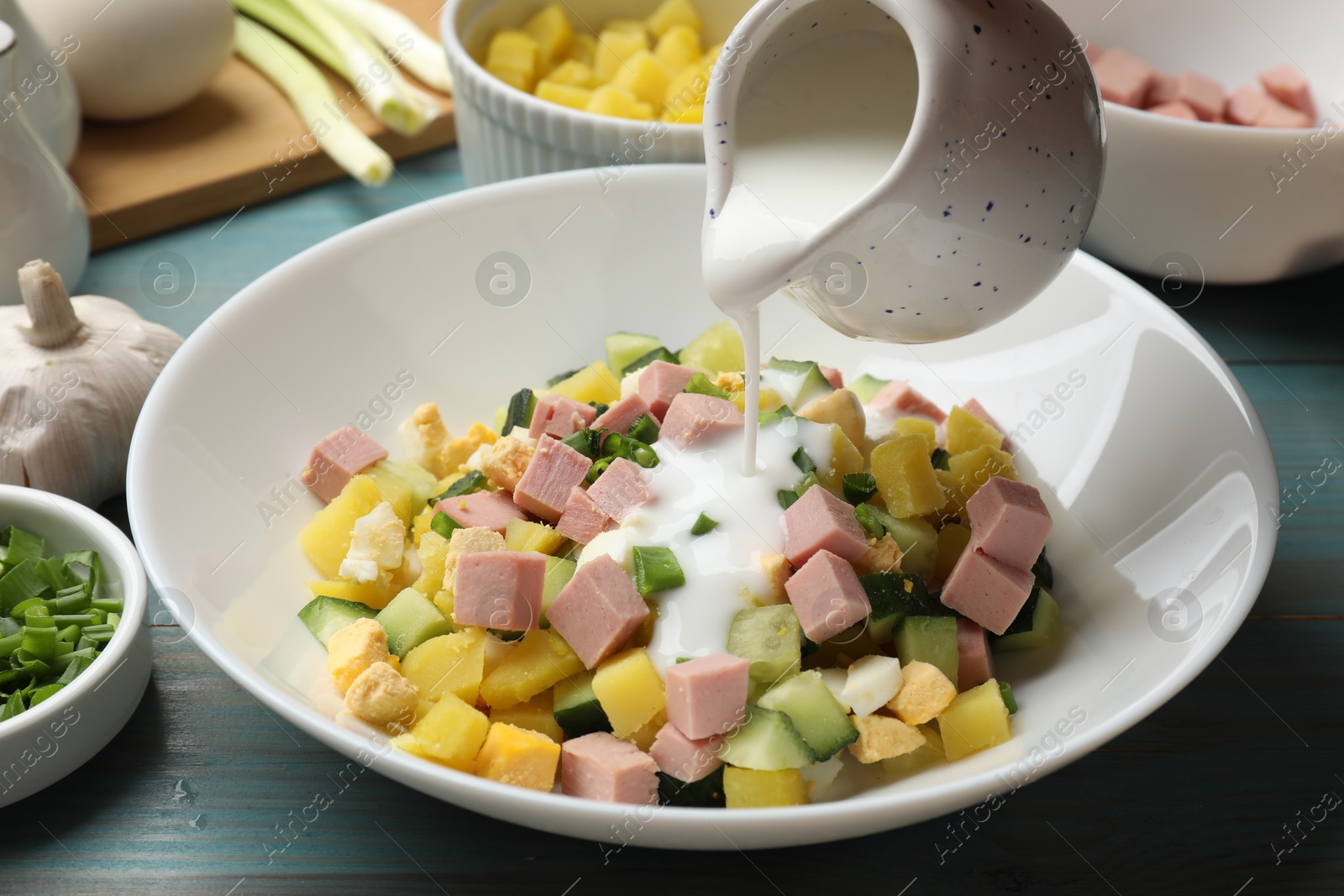 Photo of Making okroshka soup. Pouring kefir into bowl with different ingredients at light blue wooden table, closeup