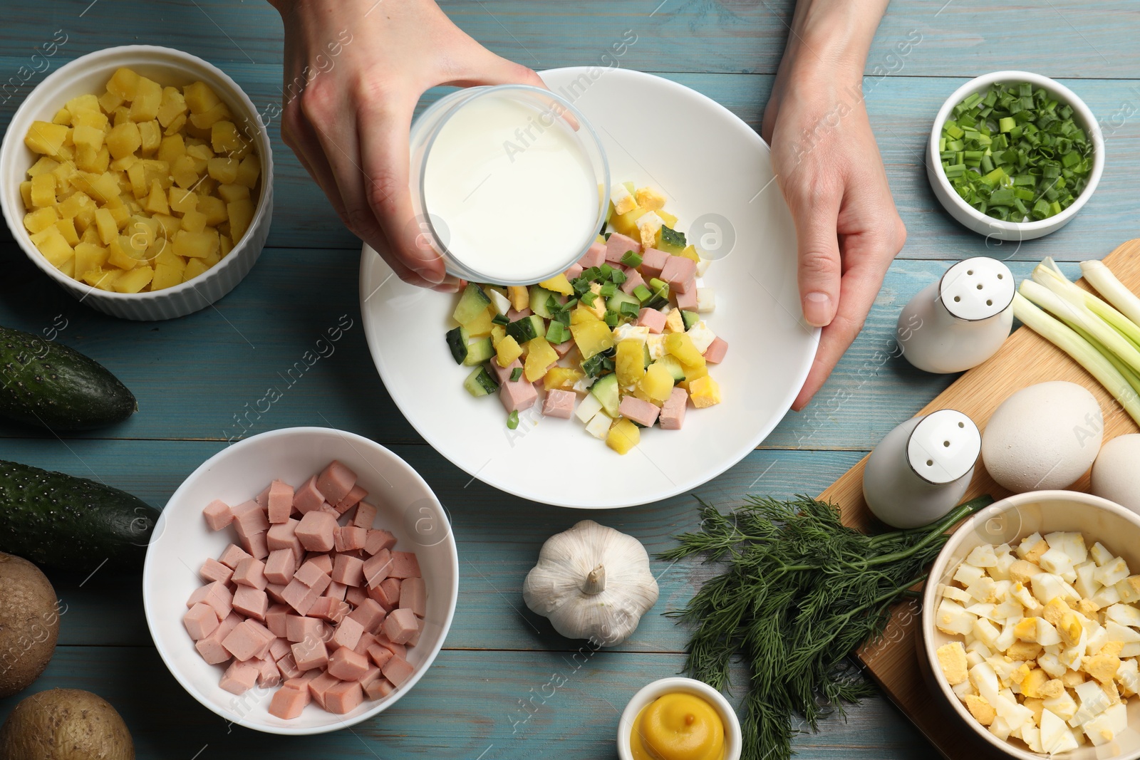 Photo of Making okroshka soup. Woman pouring kefir into bowl with different ingredients at light blue wooden table, top view