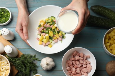 Photo of Making okroshka soup. Woman pouring kefir into bowl with different ingredients at light blue wooden table, top view