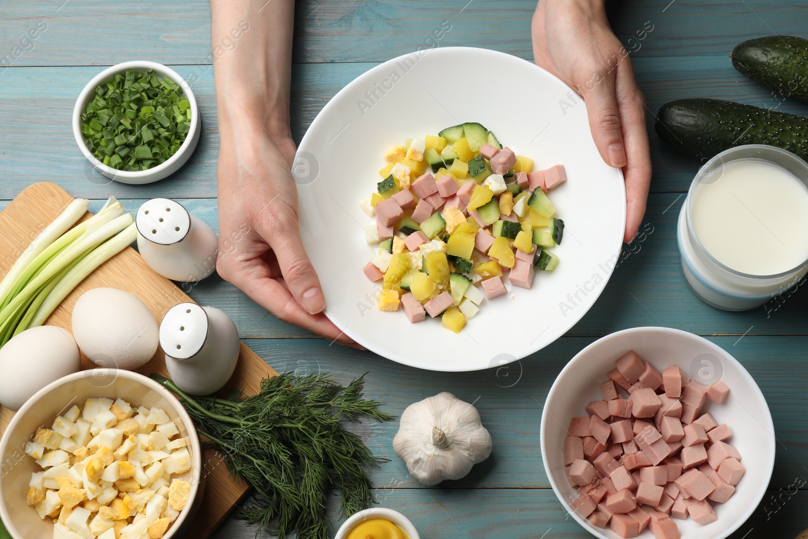 Photo of Woman with ingredients for okroshka soup at light blue wooden table, top view