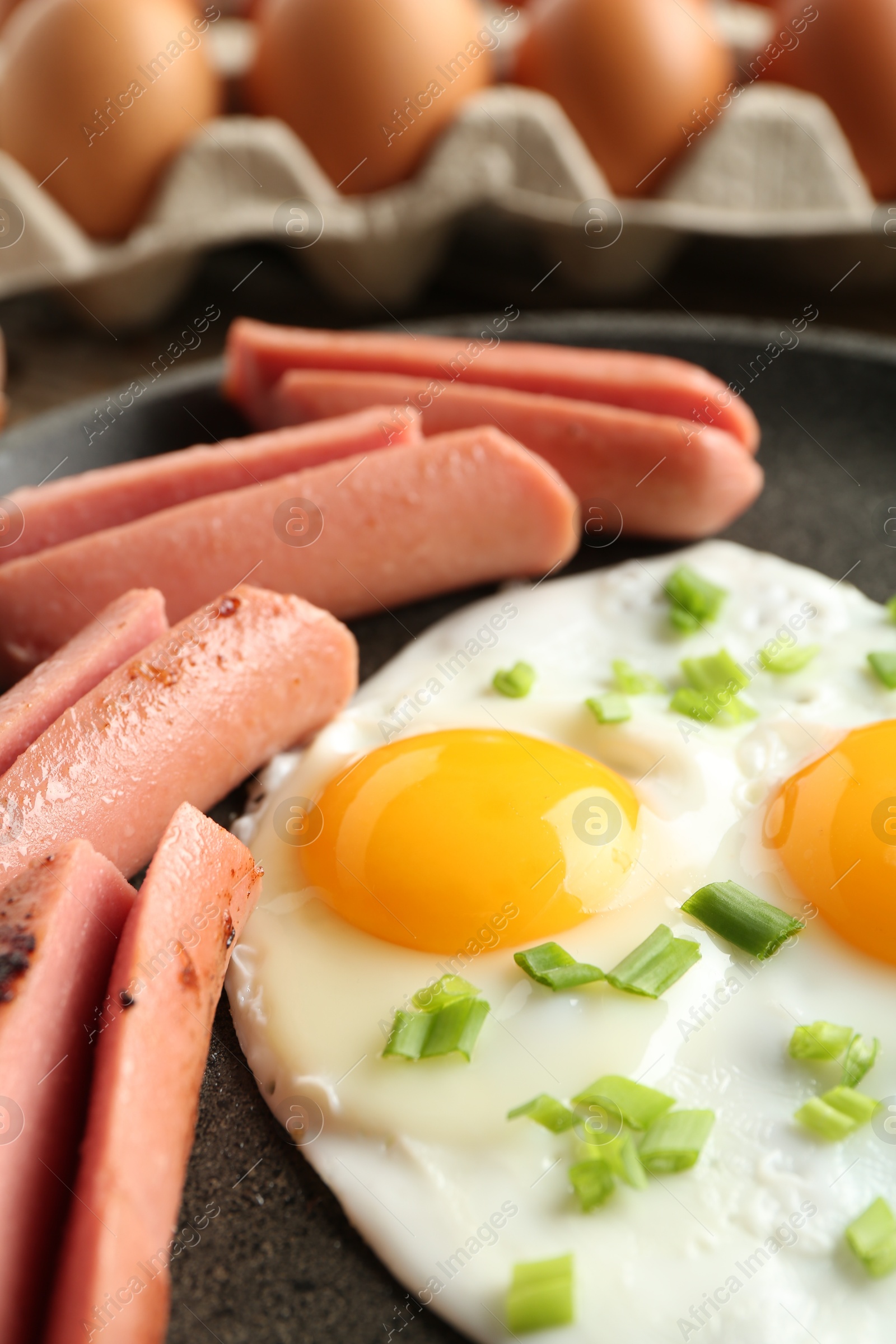 Photo of Tasty fried eggs with cut sausages and green onion in frying pan on table, closeup