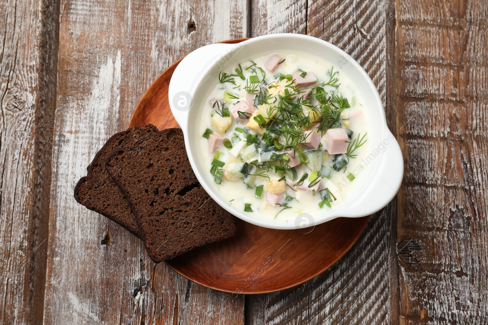 Photo of Delicious okroshka soup in bowl and bread on wooden table, top view