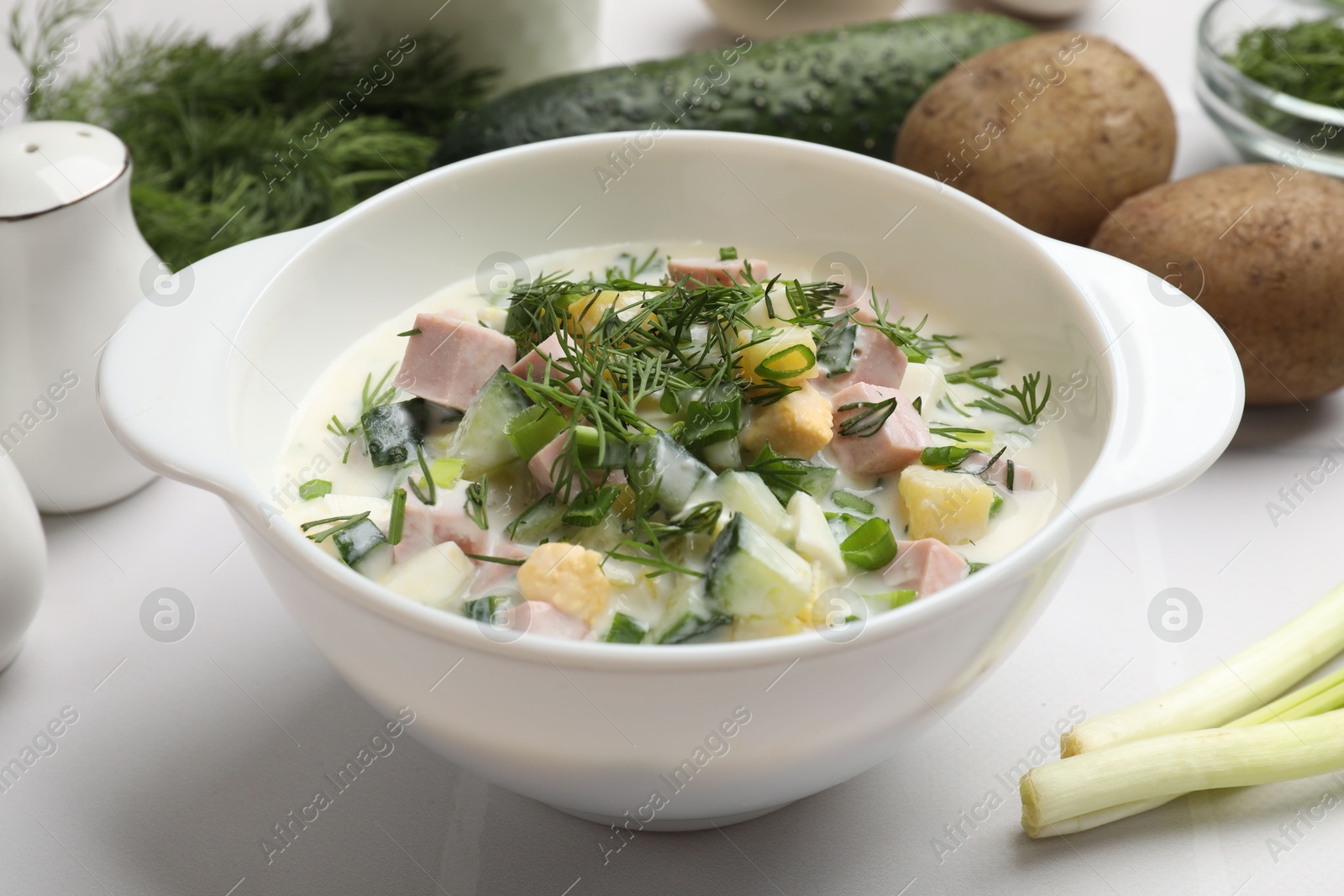 Photo of Delicious okroshka soup in bowl and ingredients on white table, closeup