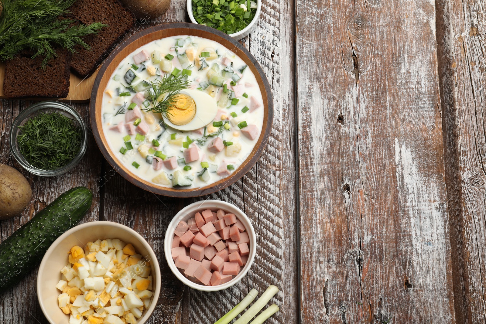 Photo of Delicious okroshka soup with kefir in bowl and ingredients on wooden table, flat lay. Space for text