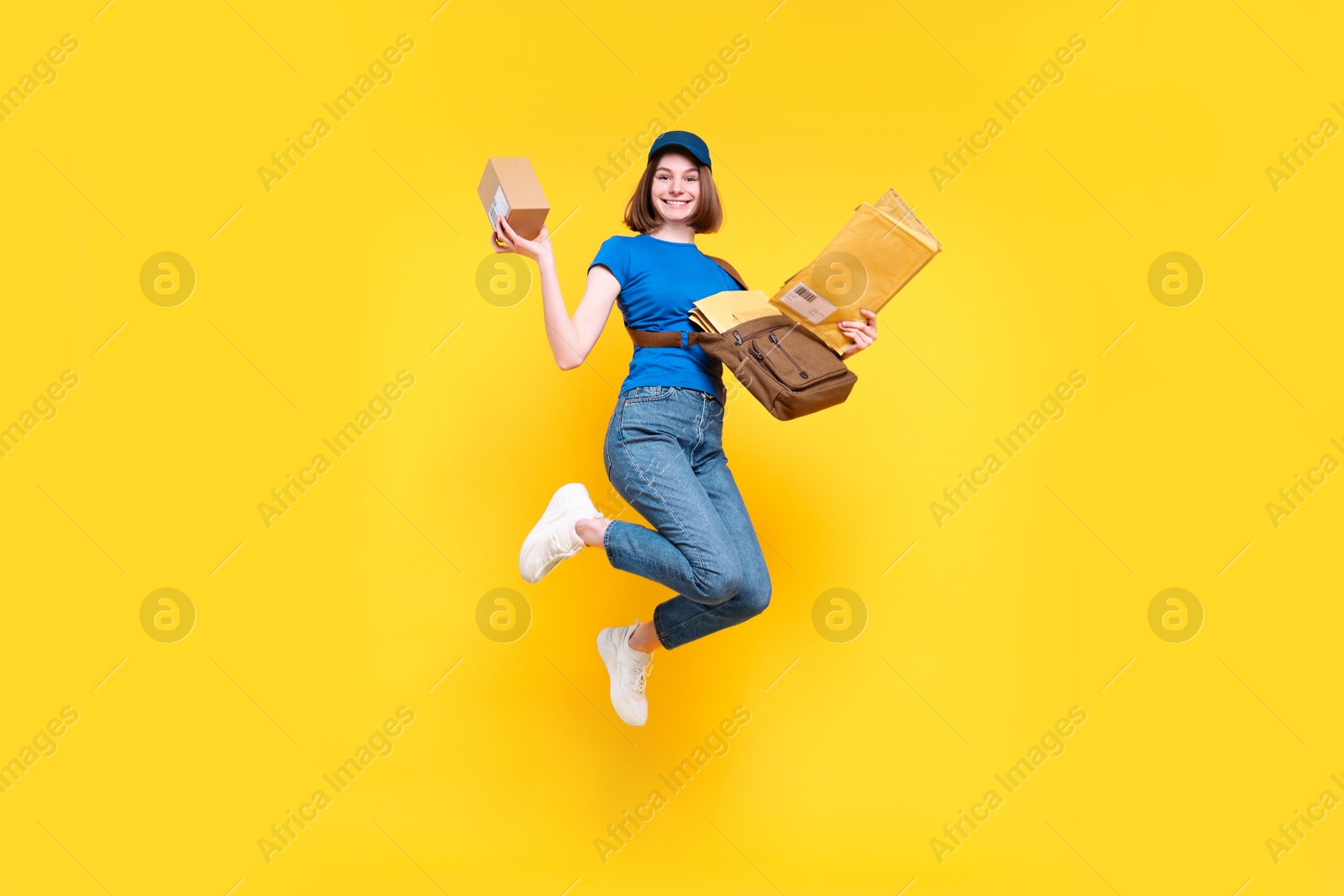 Photo of Happy postwoman with bag, parcel and envelopes jumping on yellow background