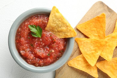 Delicious spicy salsa sauce in bowl and nacho chips on white textured table, flat lay