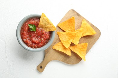 Delicious spicy salsa sauce in bowl and nacho chips on white textured table, flat lay