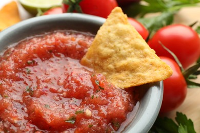 Photo of Delicious spicy salsa sauce with nacho chip in bowl and ingredients on table, closeup