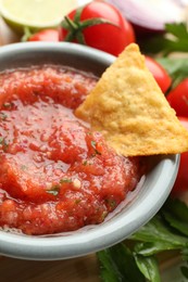 Delicious spicy salsa sauce with nacho chip in bowl and ingredients on wooden table, closeup