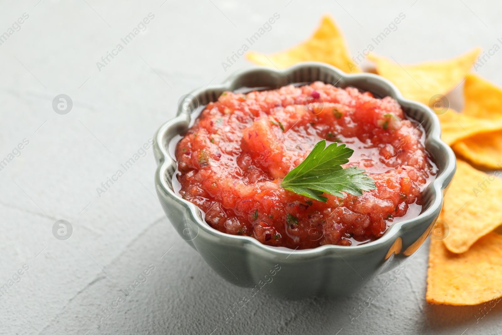 Photo of Delicious spicy salsa sauce in bowl and nacho chips on grey textured table, closeup. Space for text