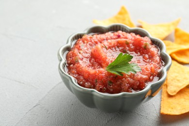 Delicious spicy salsa sauce in bowl and nacho chips on grey textured table, closeup. Space for text