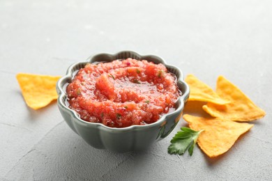 Photo of Delicious spicy salsa sauce in bowl and nacho chips on grey textured table, closeup