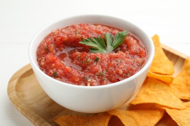 Delicious spicy salsa sauce in bowl and nacho chips on white wooden table, closeup