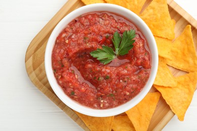 Delicious spicy salsa sauce in bowl and nacho chips on white wooden table, top view