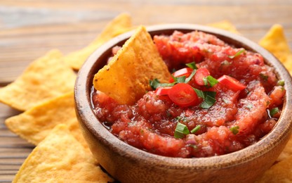 Delicious spicy salsa sauce in bowl and nacho chips on wooden table, closeup