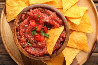 Delicious spicy salsa sauce in bowl and nacho chips on wooden table, top view