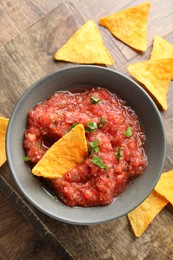 Photo of Delicious spicy salsa sauce in bowl and nacho chips on wooden table, flat lay