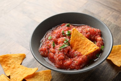 Delicious spicy salsa sauce in bowl and nacho chips on wooden table, closeup