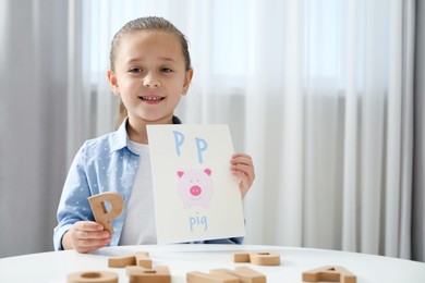 Photo of Learning alphabet. Little girl with wooden letter P and picture at white table indoors, space for text