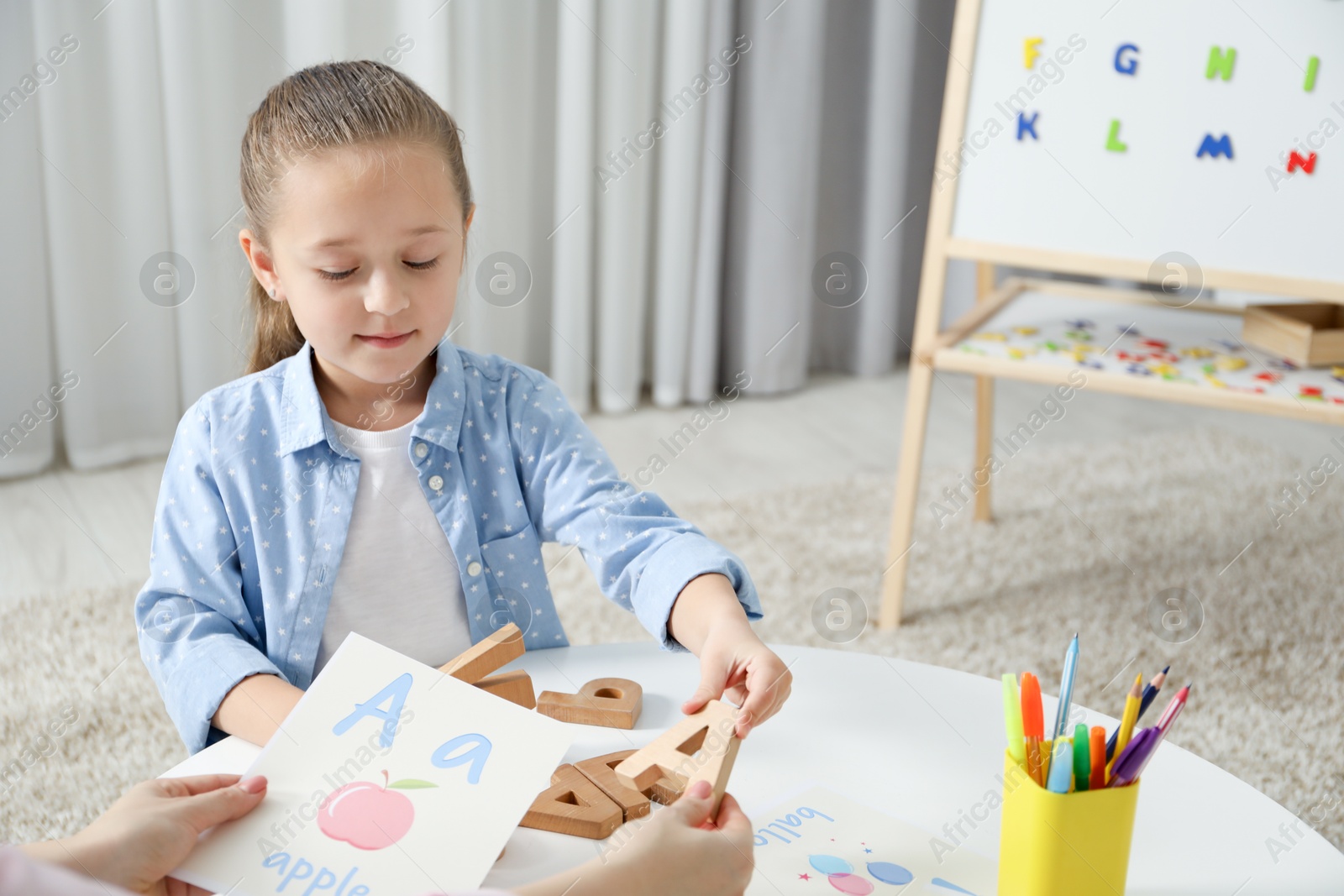 Photo of Speech therapist teaching little girl alphabet at white table indoors, closeup