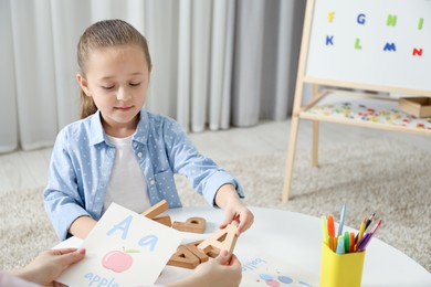 Photo of Speech therapist teaching little girl alphabet at white table indoors, closeup