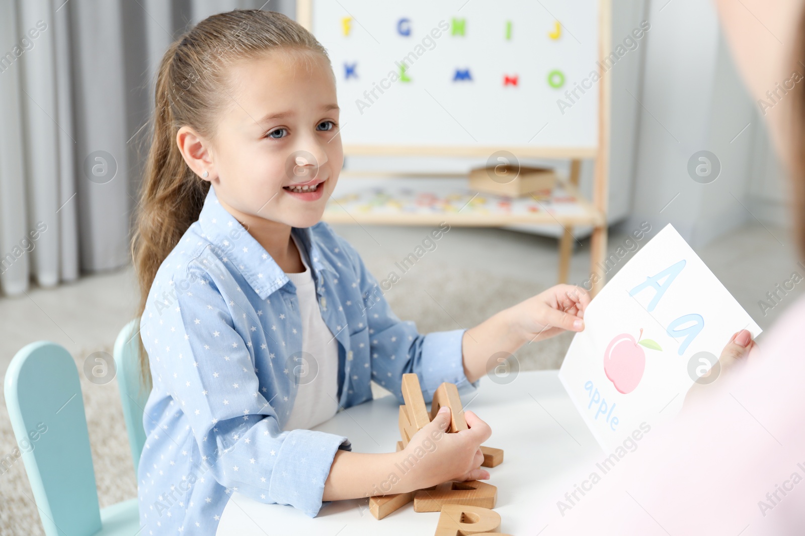 Photo of Speech therapist teaching little girl alphabet at white table indoors, closeup