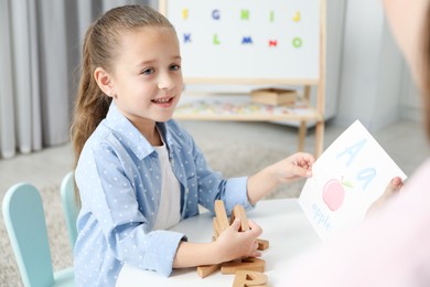 Photo of Speech therapist teaching little girl alphabet at white table indoors, closeup