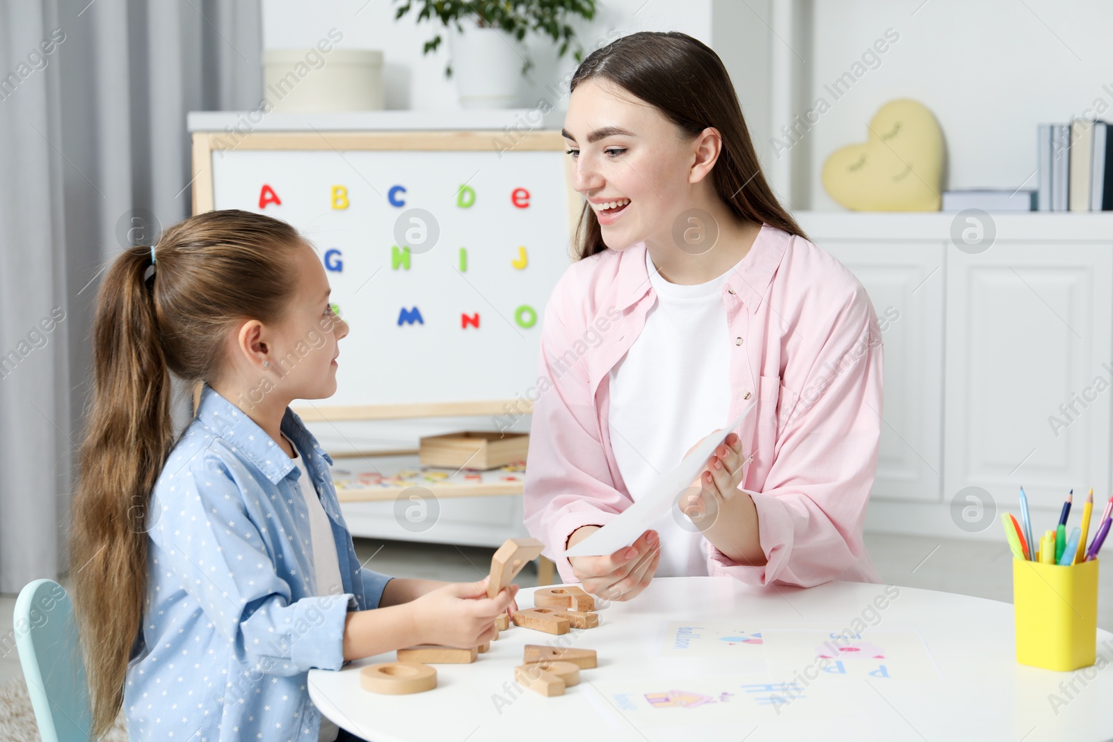 Photo of Speech therapist teaching little girl alphabet at white table indoors