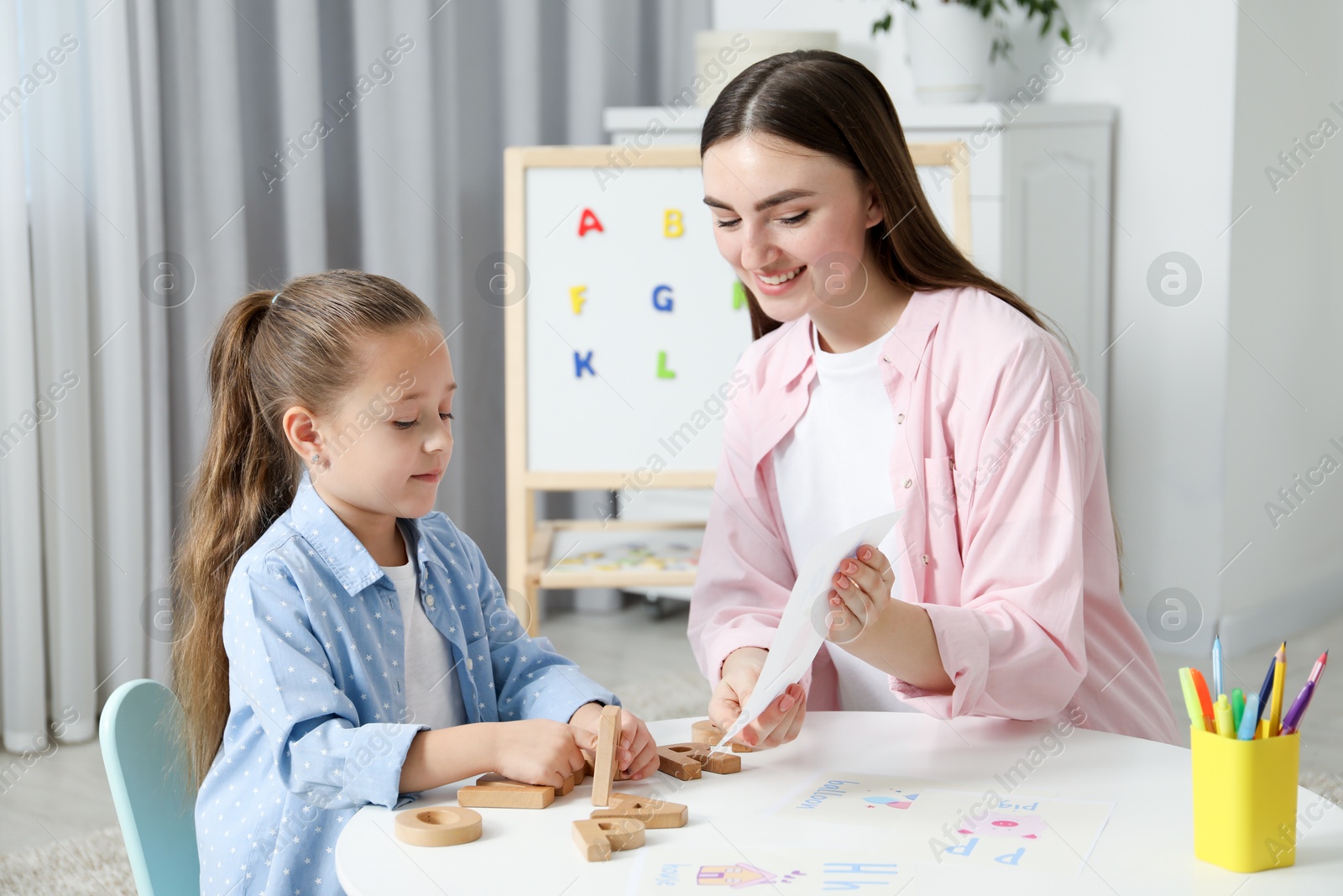 Photo of Speech therapist teaching little girl alphabet at white table indoors