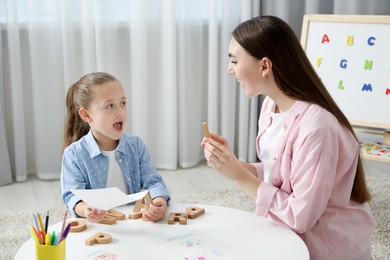 Photo of Speech therapist teaching little girl alphabet with wooden letters at white table indoors