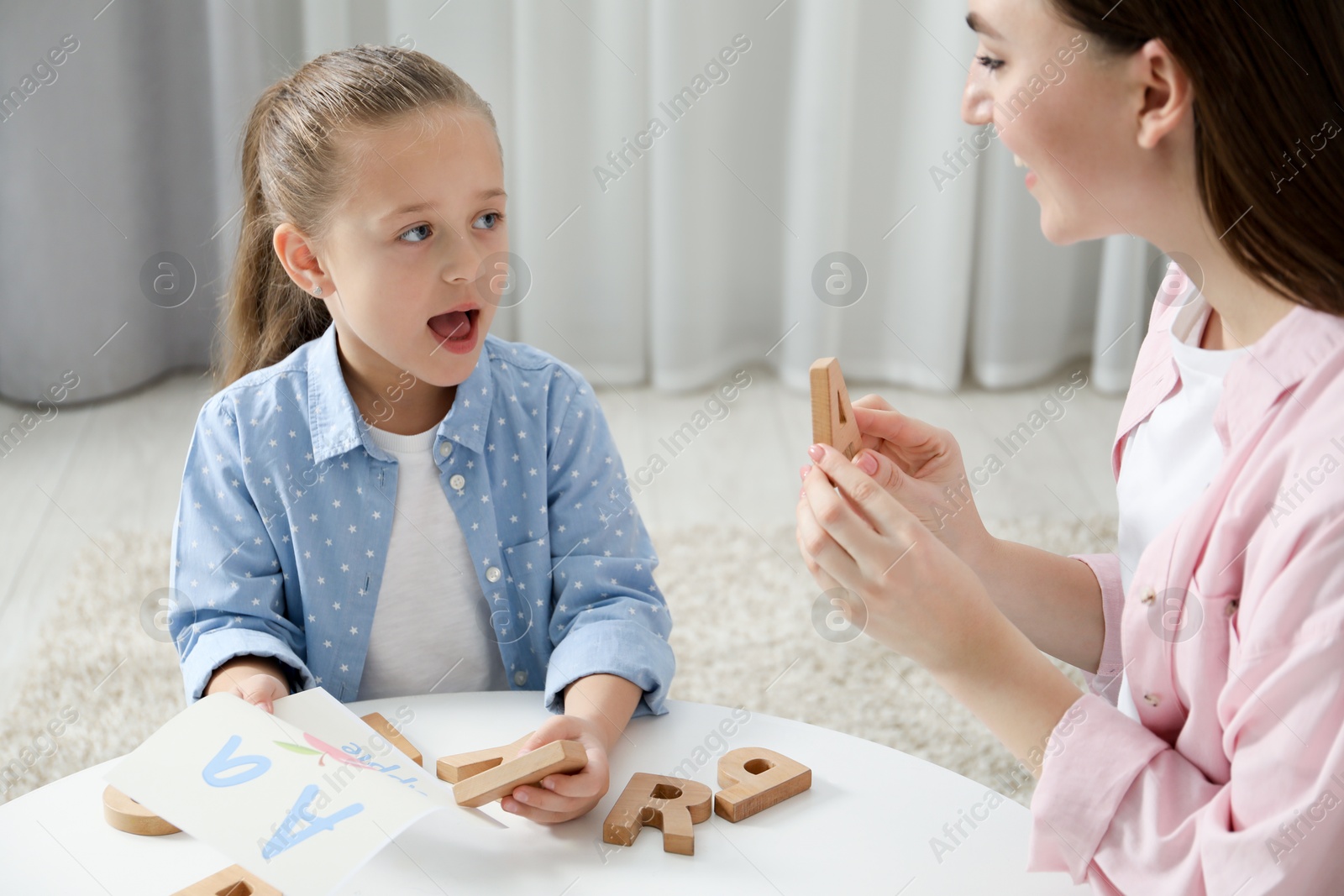 Photo of Speech therapist teaching little girl alphabet with wooden letters at white table indoors