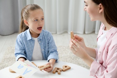 Photo of Speech therapist teaching little girl alphabet with wooden letters at white table indoors