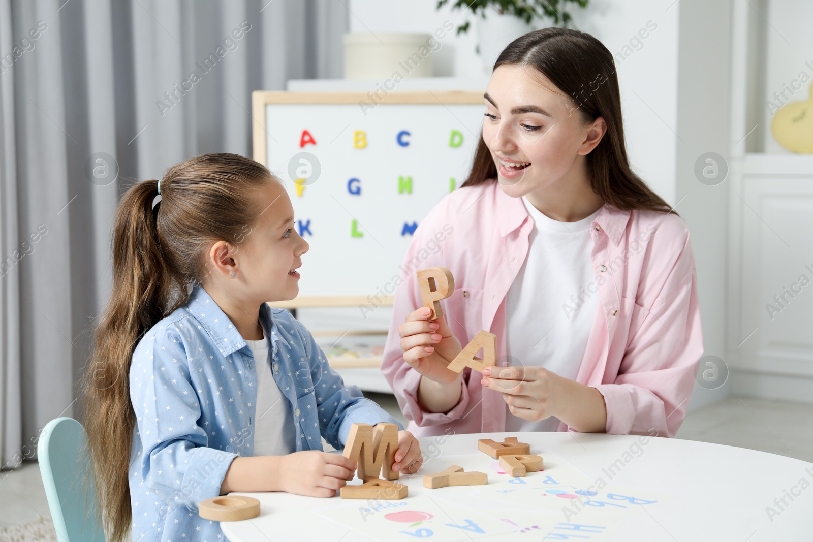 Photo of Speech therapist teaching little girl alphabet with wooden letters at white table indoors