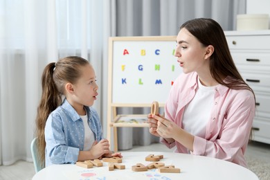 Photo of Speech therapist teaching little girl alphabet with wooden letters at white table indoors