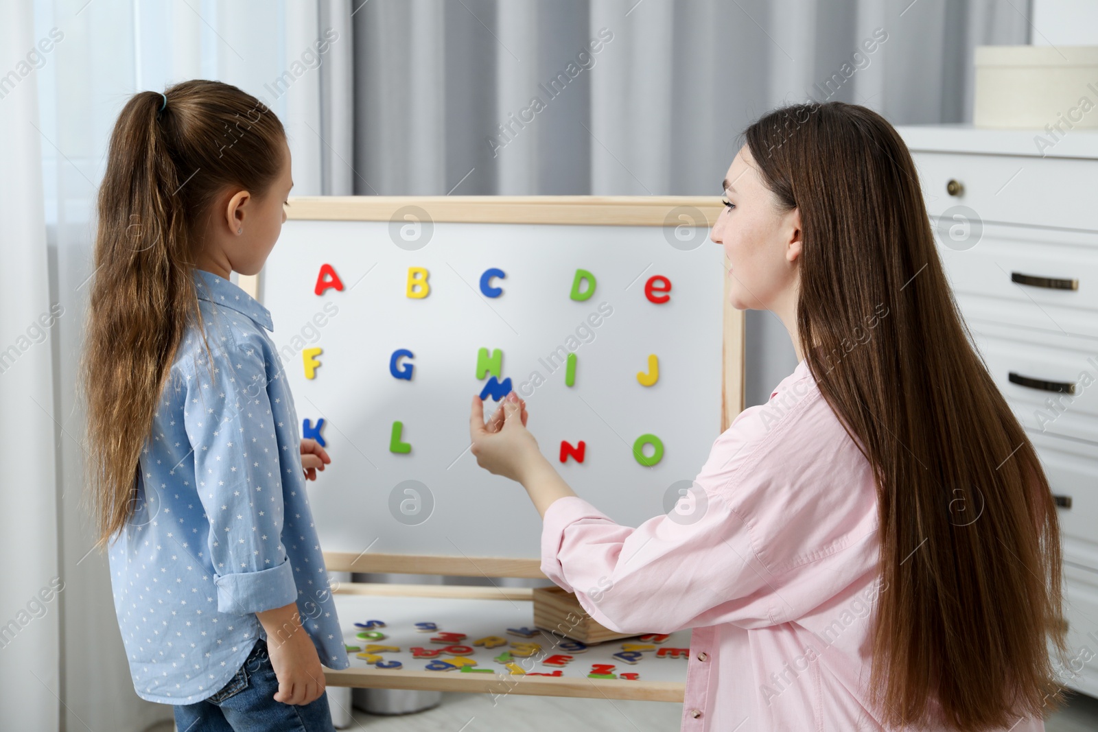 Photo of Speech therapist teaching little girl alphabet with magnetic letters indoors