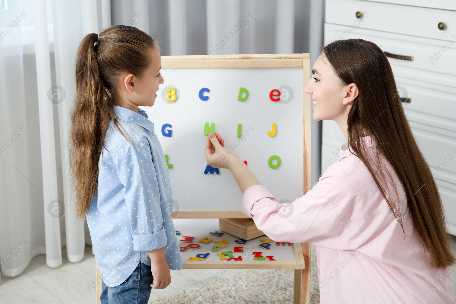 Photo of Speech therapist teaching little girl alphabet with magnetic letters indoors