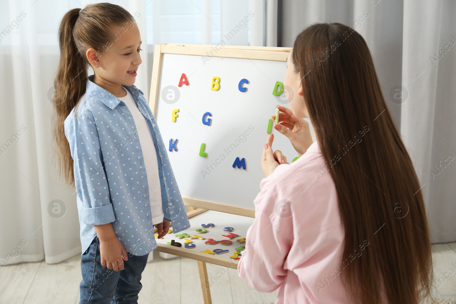 Photo of Speech therapist teaching little girl alphabet with magnetic letters indoors