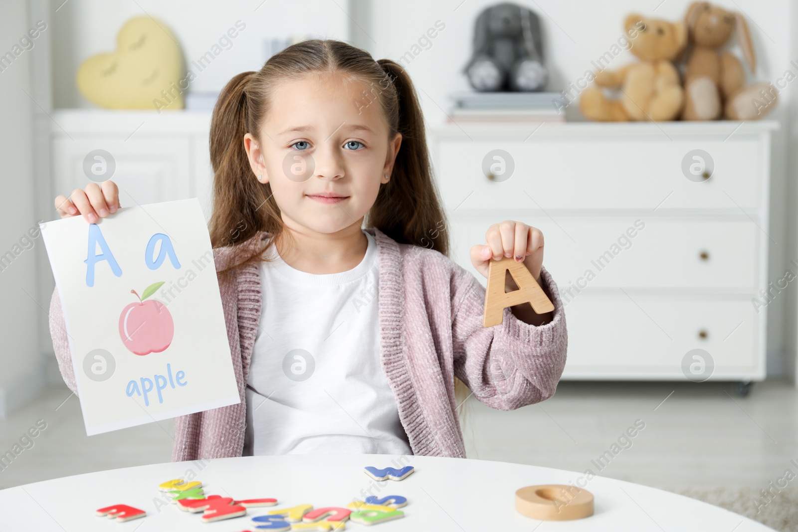 Photo of Little girl learning alphabet with different letters and picture at white table indoors