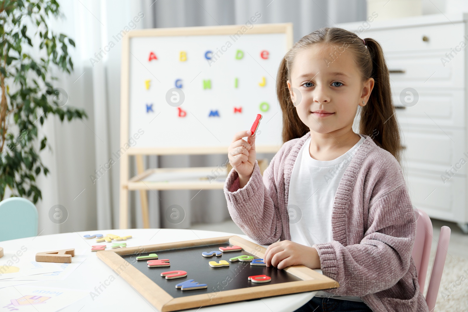 Photo of Little girl learning alphabet with magnetic letters at white table indoors