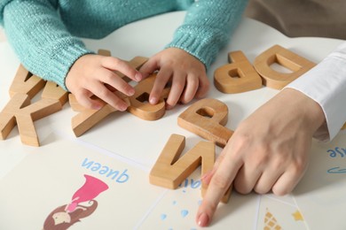 Photo of Speech therapist teaching little girl alphabet at white table indoors, closeup