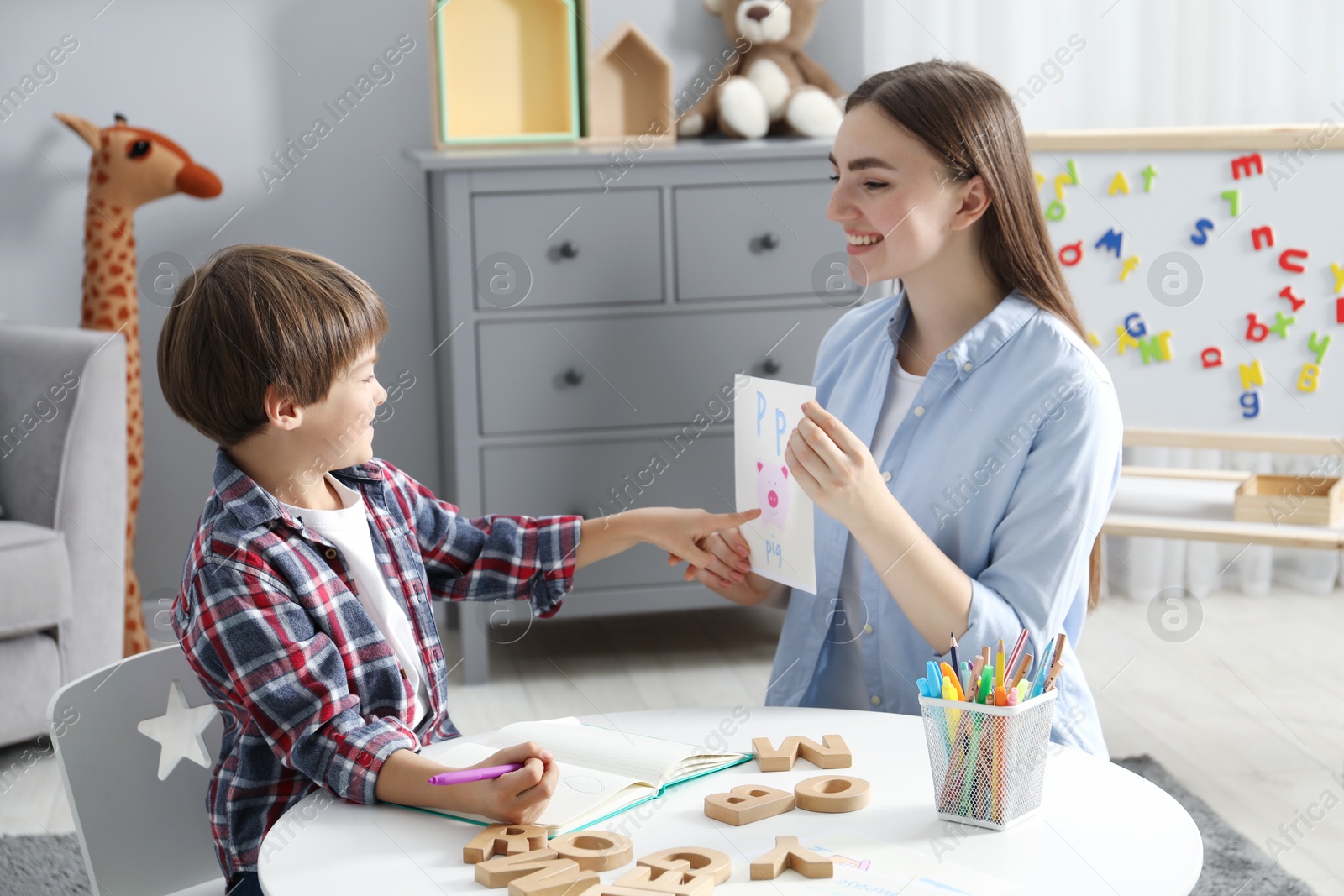 Photo of Speech therapist teaching little boy alphabet at white table indoors