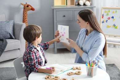 Photo of Speech therapist teaching little boy alphabet at white table indoors