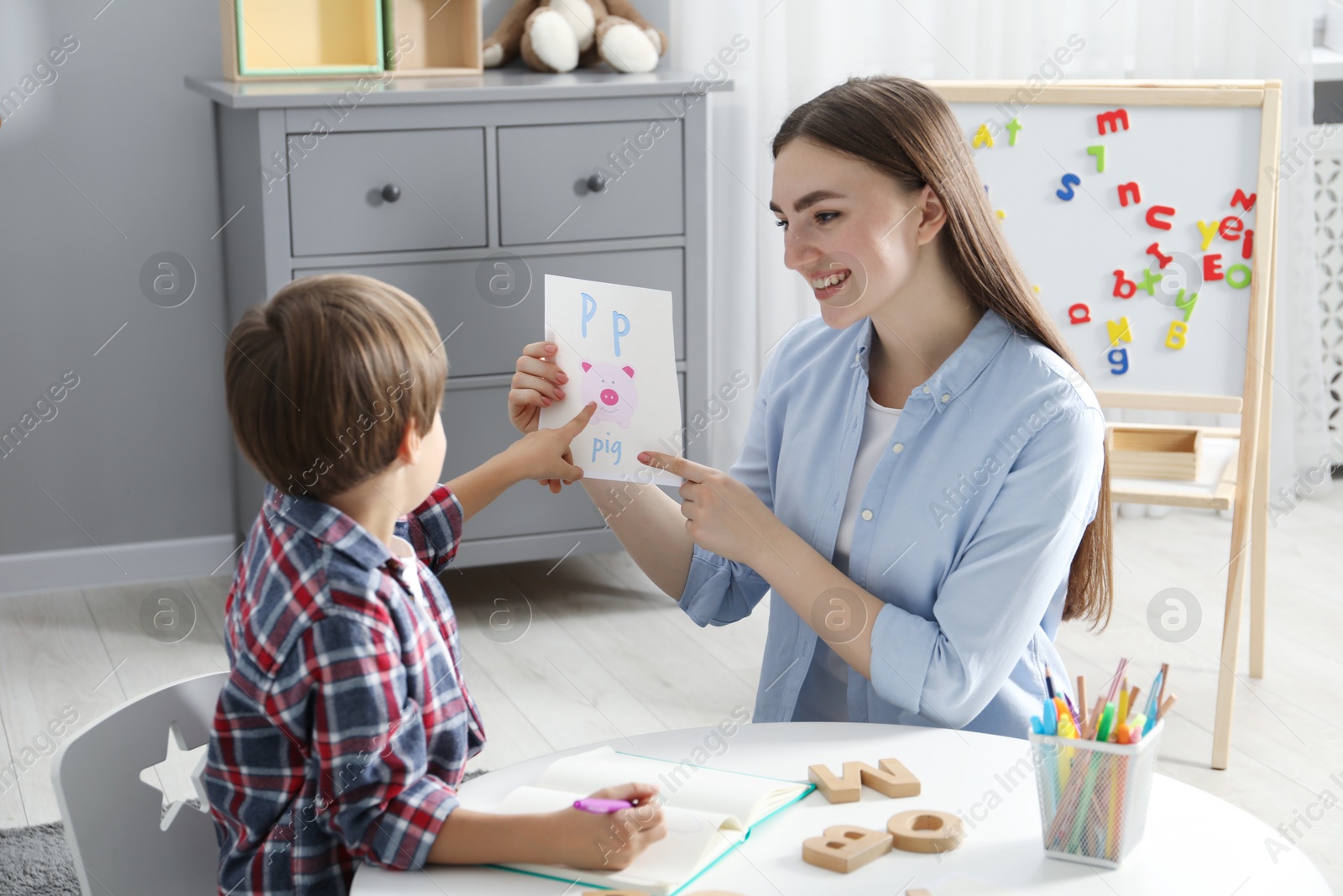 Photo of Speech therapist teaching little boy alphabet at white table indoors