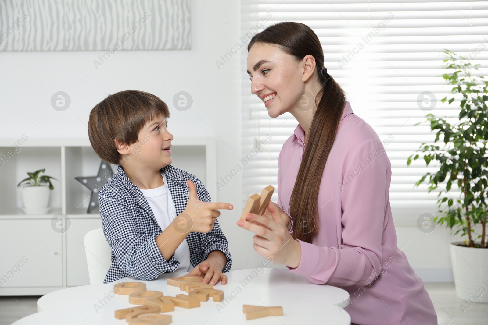 Photo of Speech therapist teaching little boy alphabet with wooden letters at white table indoors